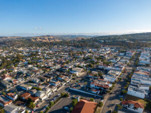 Aerial view of a suburban neighborhood with rows of houses, streets, and some hills in the background under a clear blue sky, showing an area that could be vulnerable to wildfire due to its proximity to natural landscapes.