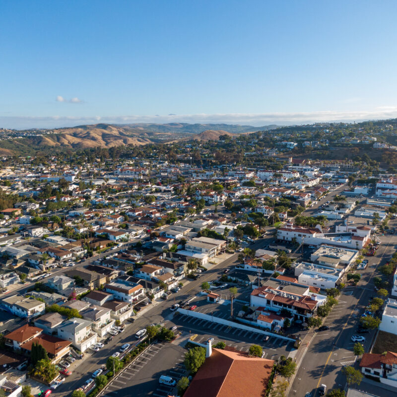 Aerial view of a suburban neighborhood with rows of houses, streets, and some hills in the background under a clear blue sky, showing an area that could be vulnerable to wildfire due to its proximity to natural landscapes.