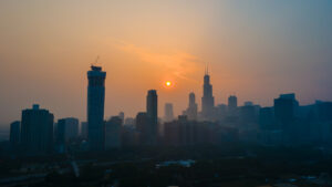 Silhouetted city skyline at sunset with the sun low in the hazy sky and tall buildings, including one under construction, against an orange and blue gradient background, as wildfire smoke drifts softly through the scene.