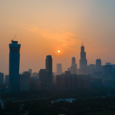 Silhouetted city skyline at sunset with the sun low in the hazy sky and tall buildings, including one under construction, against an orange and blue gradient background, as wildfire smoke drifts softly through the scene.