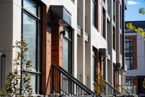 Modern apartment building exterior with large windows, wooden doors, metal railings, and small trees lining the sidewalk in bright sunlight, designed with materials chosen for added resilience against wildfire risks.