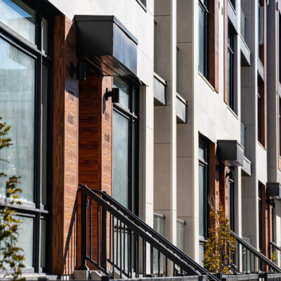Modern apartment building exterior with large windows, wooden doors, metal railings, and small trees lining the sidewalk in bright sunlight, designed with materials chosen for added resilience against wildfire risks.