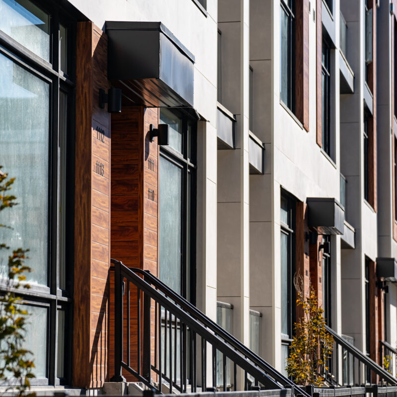 Modern apartment building exterior with large windows, wooden doors, metal railings, and small trees lining the sidewalk in bright sunlight, designed with materials chosen for added resilience against wildfire risks.