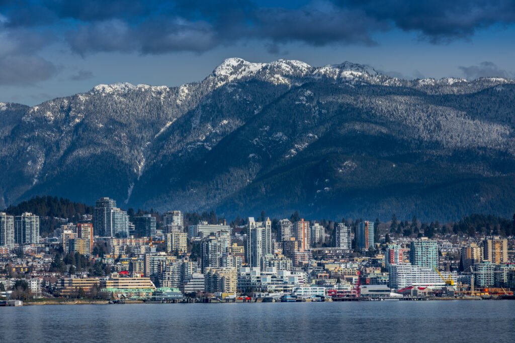 A city skyline with tall buildings stands by the water, backed by forested mountains with snowy peaks under a partly cloudy sky.