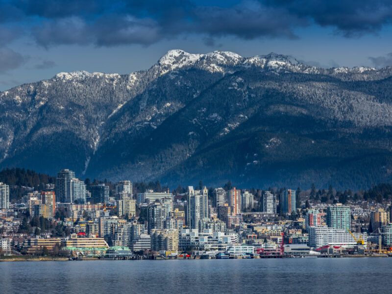 A city skyline with tall buildings stands by the water, backed by forested mountains with snowy peaks under a partly cloudy sky.