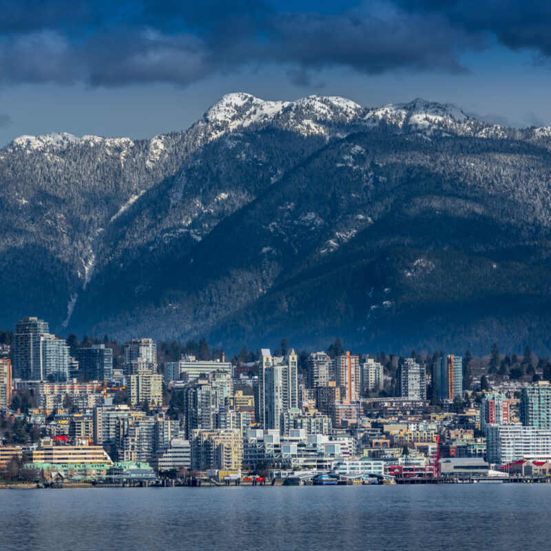 A city skyline with tall buildings stands by the water, backed by forested mountains with snowy peaks under a partly cloudy sky.