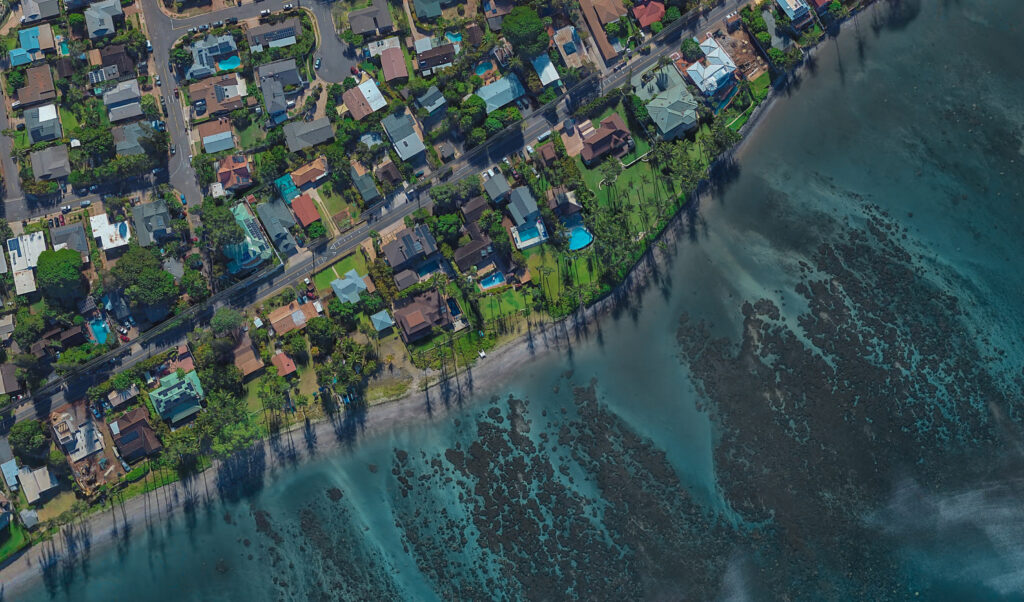 Aerial view of coastal houses bordered by a road and green lawns, with a shoreline and shallow blue ocean waters showing coral reefs or rocks below the surface—an idyllic scene untouched by wildfire.