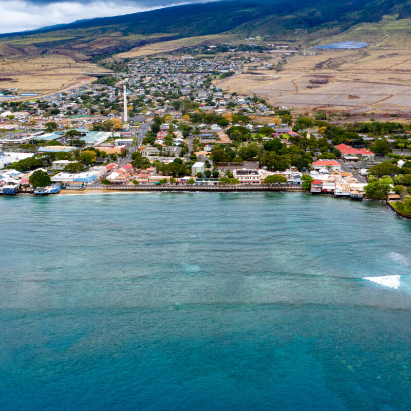 Aerial view of a coastal town with clear blue water, waterfront buildings, and green hills in the background under a cloudy sky, the landscape untouched by wildfire.