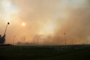 Thick smoke from a wildfire fills the sky over an empty sports field with soccer goals and tall light poles, partially blocking the sun and surrounding trees.