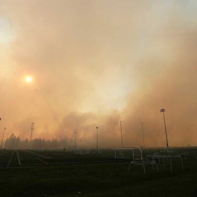 Thick smoke from a wildfire fills the sky over an empty sports field with soccer goals and tall light poles, partially blocking the sun and surrounding trees.