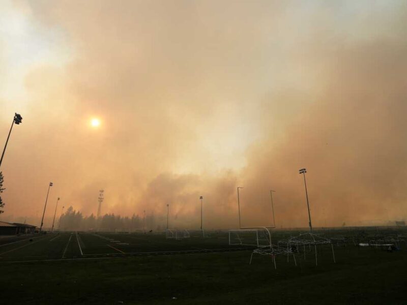 Thick smoke from a wildfire fills the sky over an empty sports field with soccer goals and tall light poles, partially blocking the sun and surrounding trees.