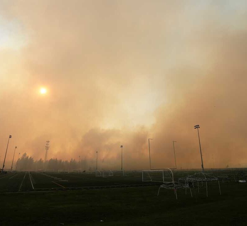 Thick smoke from a wildfire fills the sky over an empty sports field with soccer goals and tall light poles, partially blocking the sun and surrounding trees.