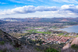 A scenic view of a sprawling suburban area near a lake, surrounded by mountains under a partly cloudy sky. Green fields and clusters of houses are visible in the foreground.