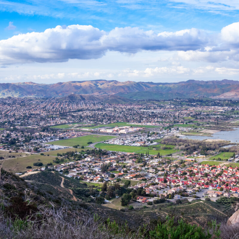 A scenic view of a sprawling suburban area near a lake, surrounded by mountains under a partly cloudy sky. Green fields and clusters of houses are visible in the foreground.