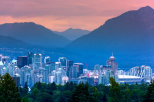Vancouver city skyline at sunset with modern buildings, BC Place Stadium, lush green trees in the foreground touched by a faint haze from distant wildfires, and mountains in the background under a colorful sky.