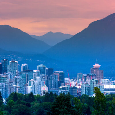Vancouver city skyline at sunset with modern buildings, BC Place Stadium, lush green trees in the foreground touched by a faint haze from distant wildfires, and mountains in the background under a colorful sky.