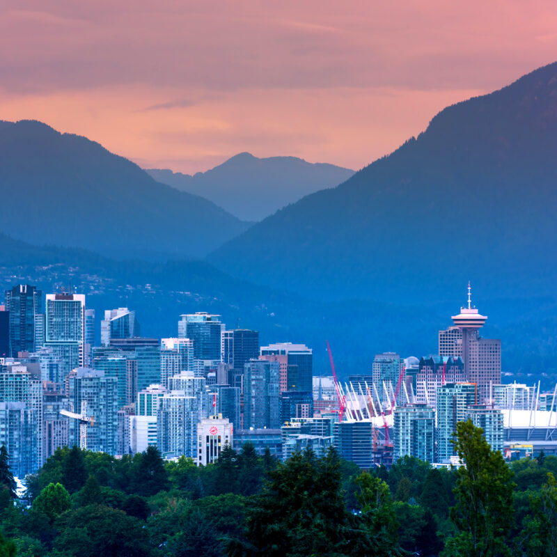 Vancouver city skyline at sunset with modern buildings, BC Place Stadium, lush green trees in the foreground touched by a faint haze from distant wildfires, and mountains in the background under a colorful sky.