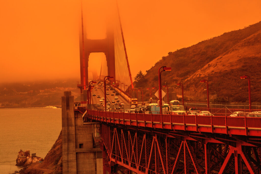 Cars cross the Golden Gate Bridge under an orange sky caused by wildfire smoke, with fog partially obscuring the towers and hills visible in the background.
