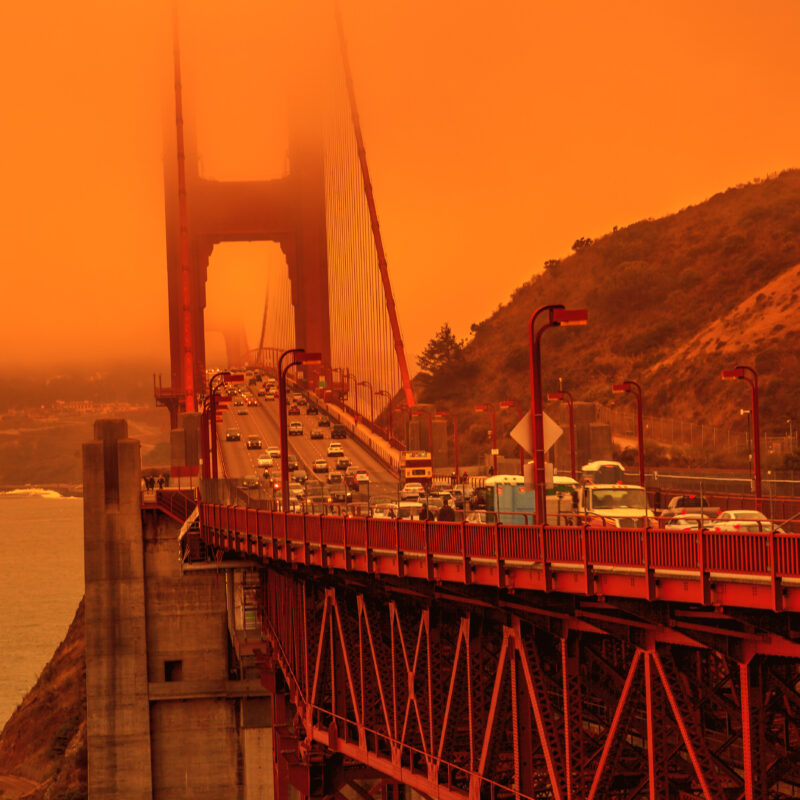 Cars cross the Golden Gate Bridge under an orange sky caused by wildfire smoke, with fog partially obscuring the towers and hills visible in the background.