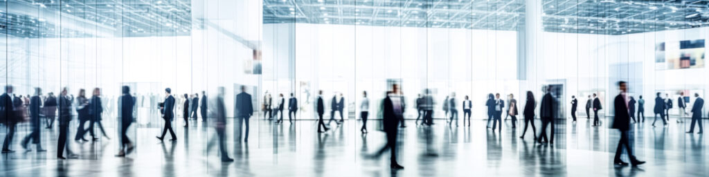 A blurred crowd of people in business attire walks through a spacious, modern, glass-walled building with high ceilings and bright lighting.