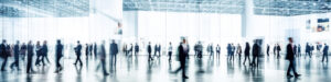 A blurred crowd of people in business attire walks through a spacious, modern, glass-walled building with high ceilings and bright lighting.