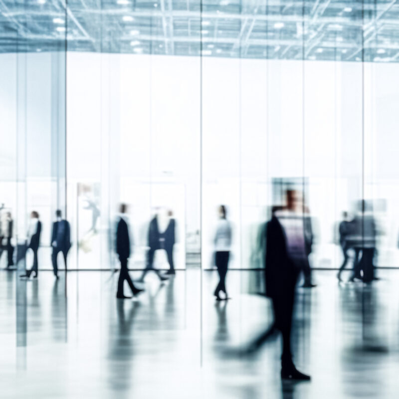 A blurred crowd of people in business attire walks through a spacious, modern, glass-walled building with high ceilings and bright lighting.