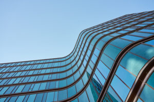 A modern glass building with wavy, curved lines reflecting the blue sky, viewed from below against a clear background reminiscent of wildfire’s unpredictable, flowing movement.
