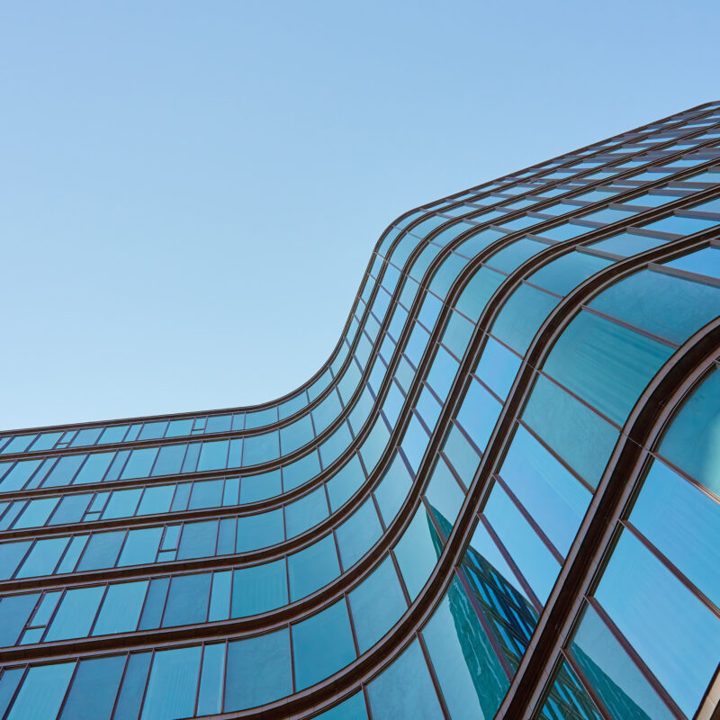 A modern glass building with wavy, curved lines reflecting the blue sky, viewed from below against a clear background reminiscent of wildfire’s unpredictable, flowing movement.