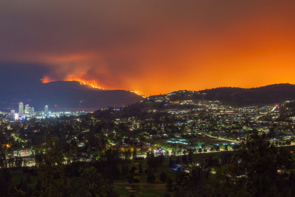 A city at night sits below a mountainside engulfed in a wildfire, with an intense orange glow filling the sky.