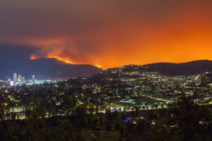 A city at night sits below a mountainside engulfed in a wildfire, with an intense orange glow filling the sky.