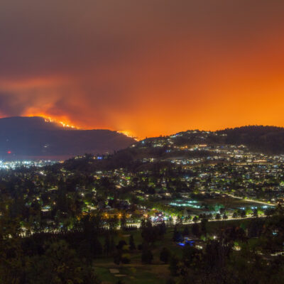 A city at night sits below a mountainside engulfed in a wildfire, with an intense orange glow filling the sky.