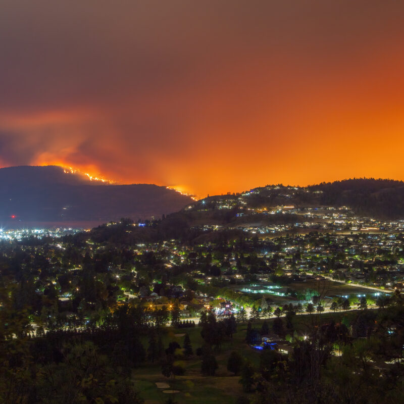 A city at night sits below a mountainside engulfed in a wildfire, with an intense orange glow filling the sky.