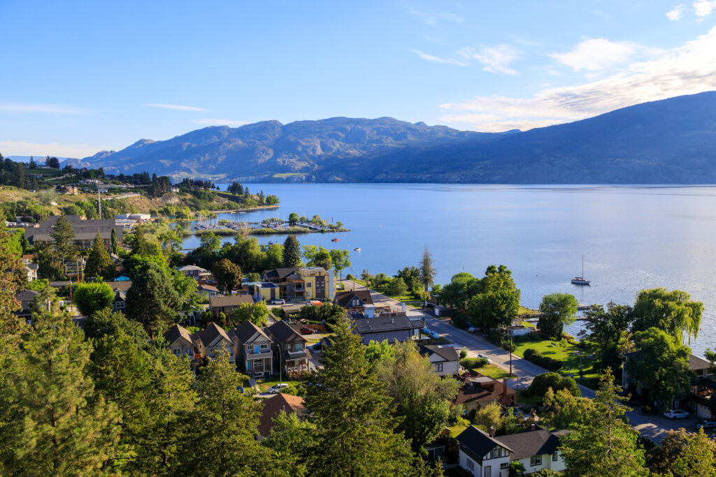 A high-angle view of a residential neighborhood along the shore of a large lake, with mountains in the background and a small marina in the distance, partially obscured by wildfire smoke.