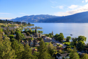 A high-angle view of a residential neighborhood along the shore of a large lake, with mountains in the background and a small marina in the distance, partially obscured by wildfire smoke.