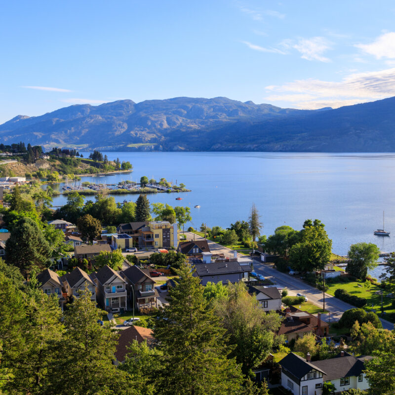 A high-angle view of a residential neighborhood along the shore of a large lake, with mountains in the background and a small marina in the distance, partially obscured by wildfire smoke.